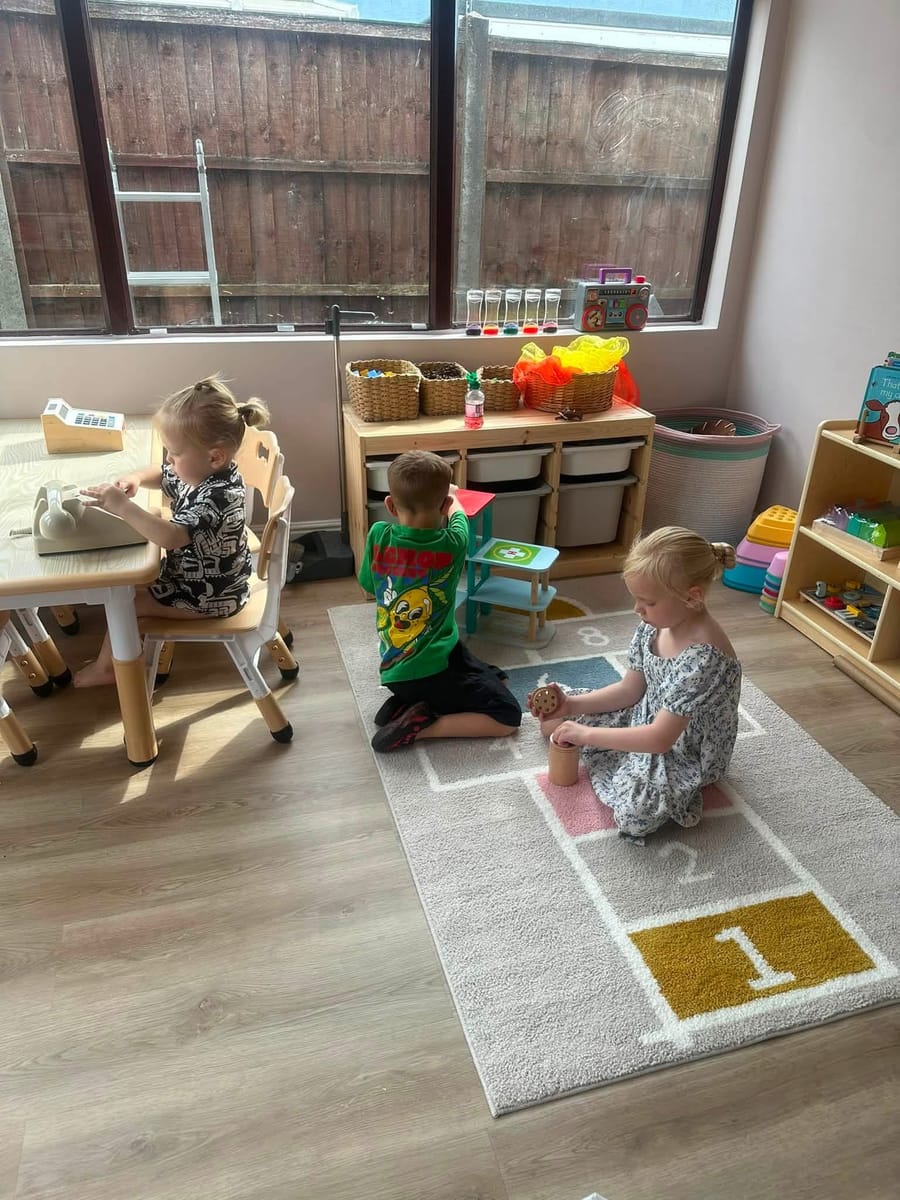 Three children playing in the bright playroom on a hopscotch rug, with wooden shelves and baskets of toys