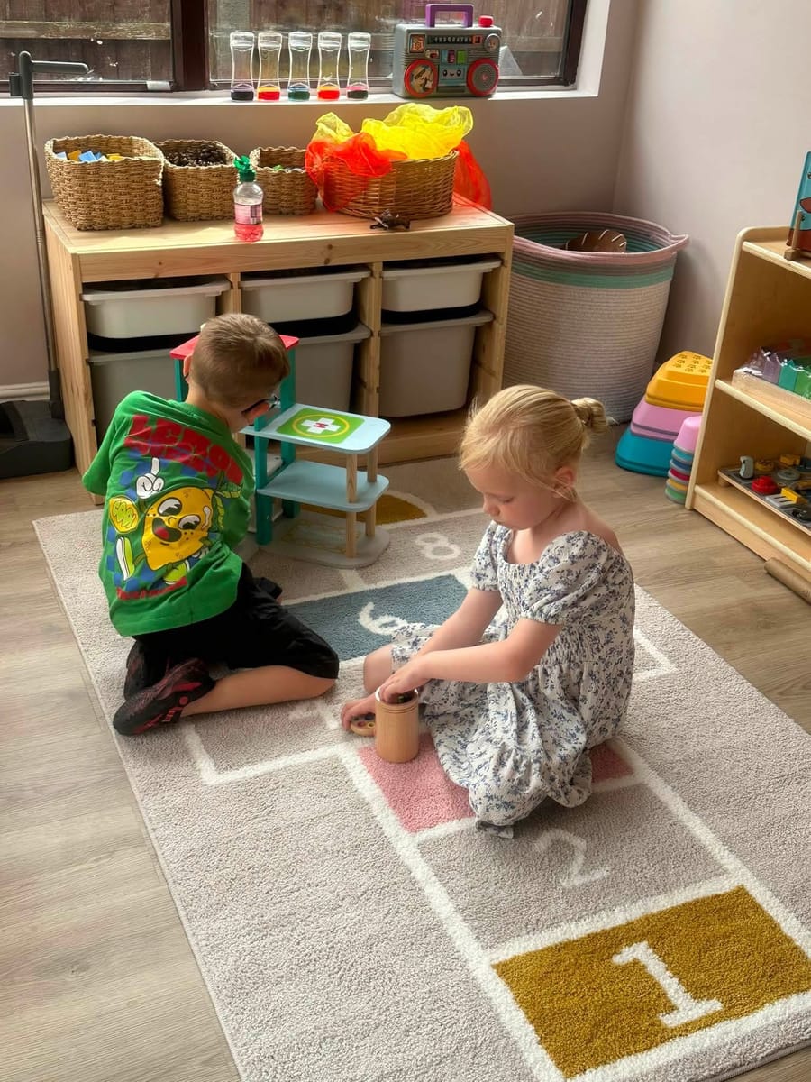 Two children sitting together on the playroom rug, exploring wooden toys side by side