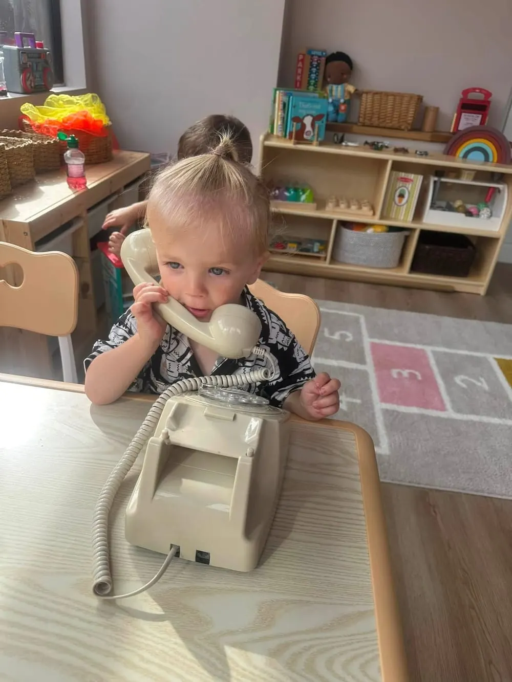A toddler chatting on a toy telephone at a low wooden table in the playroom